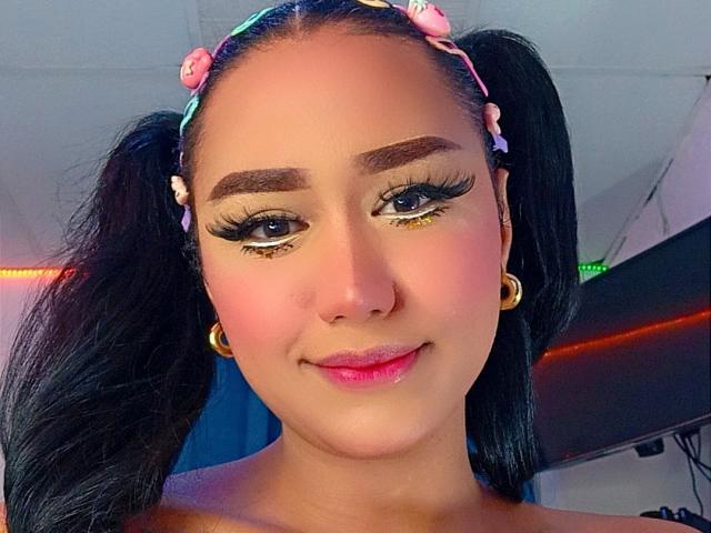 AnielFetish has dark pigtails, pink flower headband, gold hoop earrings, heavy eyeliner, glittery eyelids, pink blush, and a cute smile. She's a sexy Latina with a cute face.