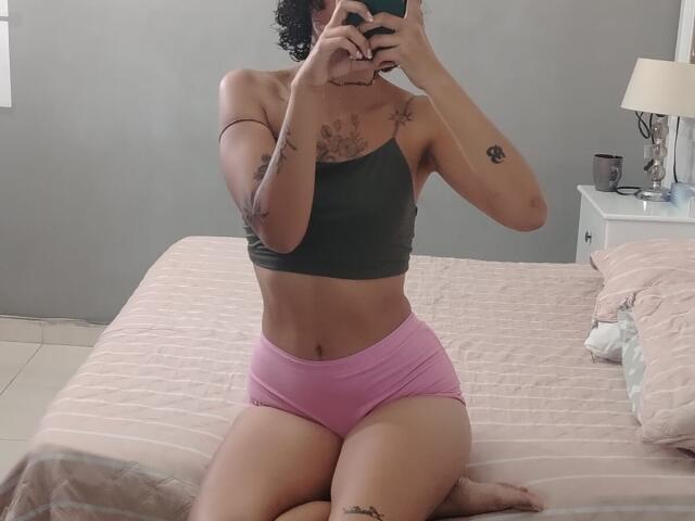 SilvanaXOwen with curly hair, wearing a green crop top and pink boyshorts, kneeling on a bed, showing tattoos on arms and chest, taking a mirror selfie.