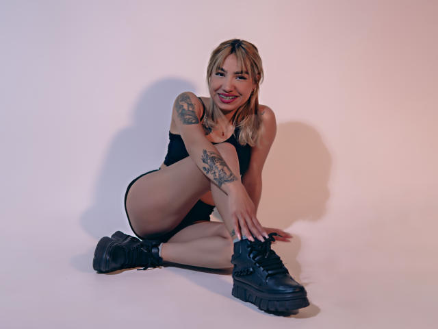AnneGela with blonde hair, black tank top, and shorts, sitting on the floor, smiling, tattoos on arm, black boots, knees up, hands on boots, sexy, cute.
