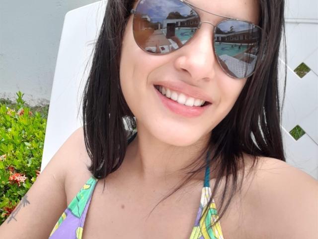 CamityTorresx with black hair, wearing reflective sunglasses, smiling, showing white teeth, in a colorful bikini top, has a small tattoo on her right arm, sitting outdoors.
