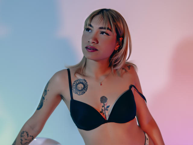 AnneGela, com cabelo loiro, sutiã preto, tatuagens no peito e nos braços, olhando para cima com lábios rosados, iluminação suave, braços ligeiramente levantados. Ela tem seios pequenos e uma tatuagem de flor..