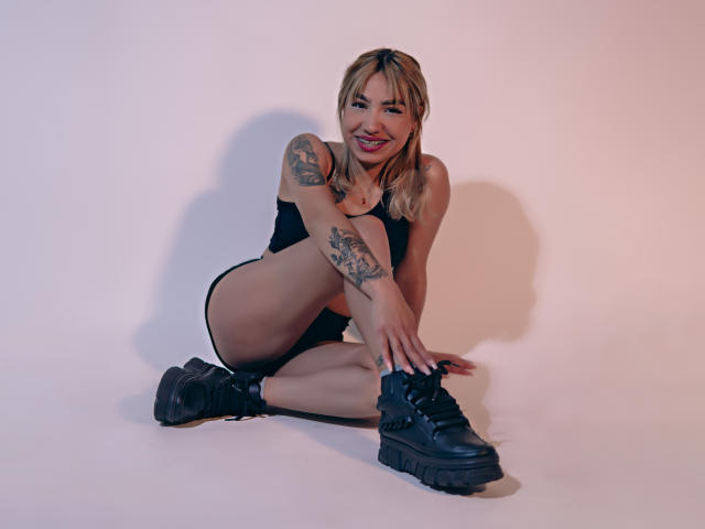AnneGela, loira com tatuagens, sentada no chão, vestindo shorts pretos e botas, sorrindo, mostrando seus seios, braços e pernas, com o bumbum ligeiramente visível, em uma foto de estúdio..