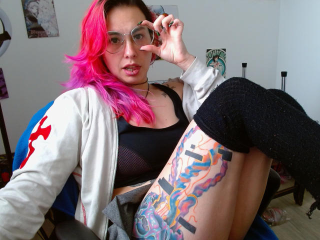 NatashaLau with pink hair, glasses, black mesh top, white jacket, black knee socks, colorful thigh tattoo, and ring, sitting in chair, showing her tits and pussy through top.