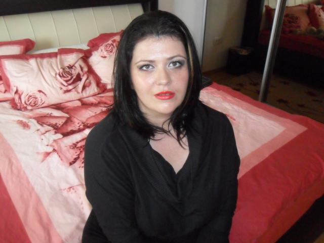 Carolina69 has pale skin, black hair, red lipstick, and smoky eye makeup, wearing a black top, sitting in front of a red and pink rose-patterned bed.