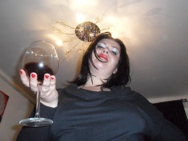 Carolina69 met rode lippenstift, een zwarte trui en rode nagellak, die een glas rode wijn vasthoudt, lachend en naar beneden kijkend in de richting van de camera, onder een plafondlamp met een sterrenpatroon..
