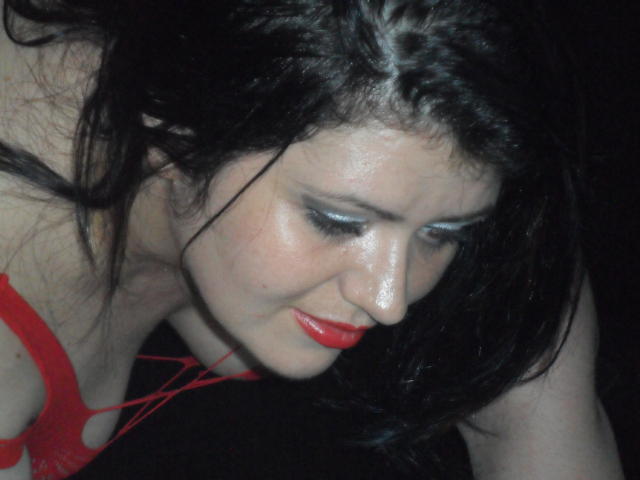 Carolina69 with black hair, red lipstick, and shiny blue eye makeup, leaning forward in a red top, showing her tits and looking down with a slightly anxious expression.