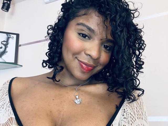 LisaMendez, avec ses cheveux noirs bouclés, sa peau brun clair, son rouge à lèvres rouge et son collier en forme de cœur, sourit à l'appareil photo, montrant sa poitrine dans un haut blanc en tricot..