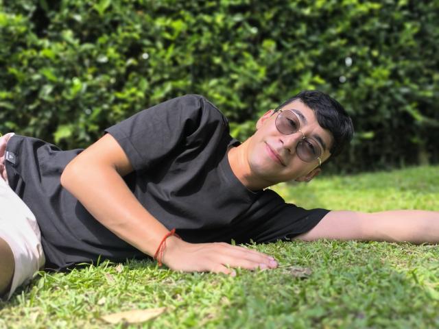 OliverHot69, deitado na grama, vestindo uma camiseta preta, shorts brancos, uma pulseira vermelha e óculos, com uma expressão relaxada, um sorriso leve e cabelo preto curto..