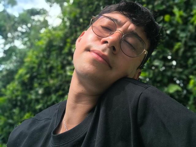 OliverHot69 with glasses, black shirt, and necklace, head tilted back, eyes closed, slight smile, short dark hair, green leafy background.