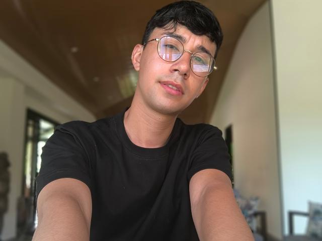OliverHot69 with short black hair, glasses, and a black t-shirt, making a slight pouty face in a selfie. His cute, slightly chubby face is in focus.