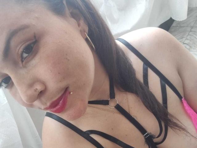 Vennus69, con i capelli castano scuro, i piercing al naso e il rossetto rosso, indossa un intimo nero con spalline sottili, una collana a catena dorata e, sul lato destro, si intravede un reggiseno rosa..