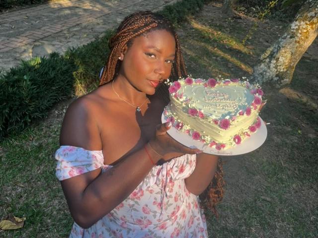 AkaneCox with dark skin, braids, and a floral dress holds a pink and white frosted cake with pink roses. Her tits are small, and she's pouting slightly.