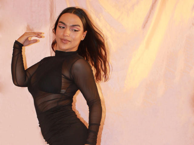 Sophie Carter, torride dans une robe noire transparente, dévoilant ses seins et ses fesses à travers le tissu, avec de longs cheveux noirs et un fard à paupières rose, debout devant un fond rosâtre..