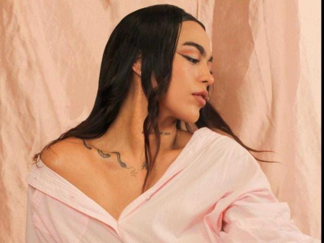 Sophie Carter : Cheveux noirs longs, chemisier rose à épaules dénudées, tatouage sur le cou, lèvres pulpeuses et maquillage mettant en valeur ses yeux, sur un fond rose clair..