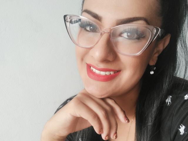 LailaRainbow has red lipstick, clear glasses, black hair, and a smile showing her teeth. Her hand rests on her chin, with her nails painted a dark red.