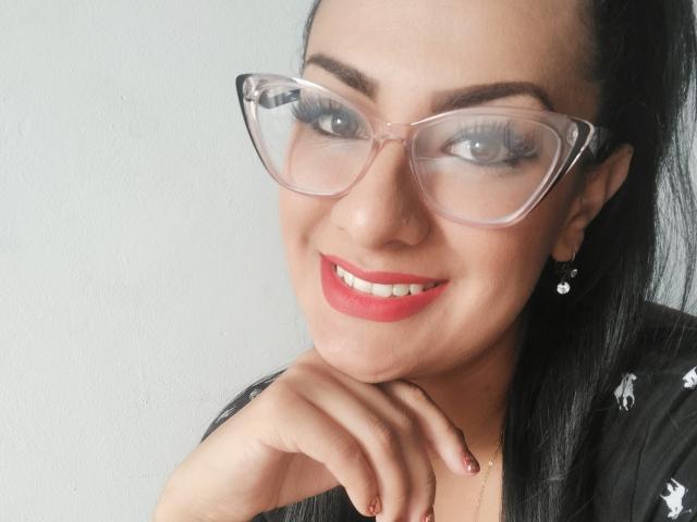 LailaRainbow mit Brille, rotem Lippenstift und schwarzem Haar, lächelnd mit der Hand am Kinn, trägt ein schwarzes Hemd, hat eine Nasenring und kleine Ohrringe..