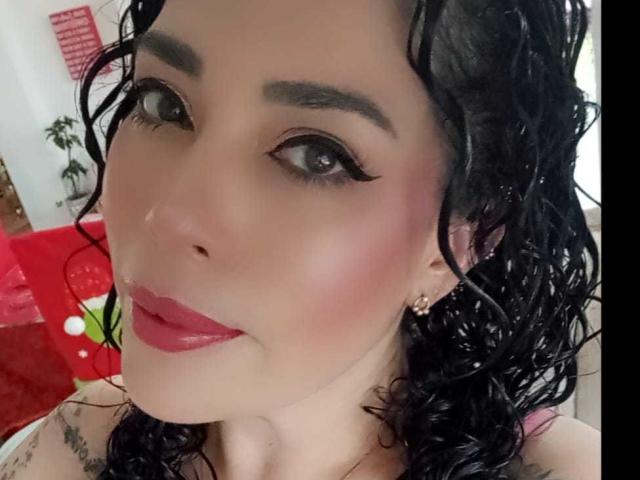 LyemDreams has curly black wet hair, dark eyebrows, eyeliner, red lipstick, and earrings, with tattoos on her neck. Her pinkish skin is slightly shiny.