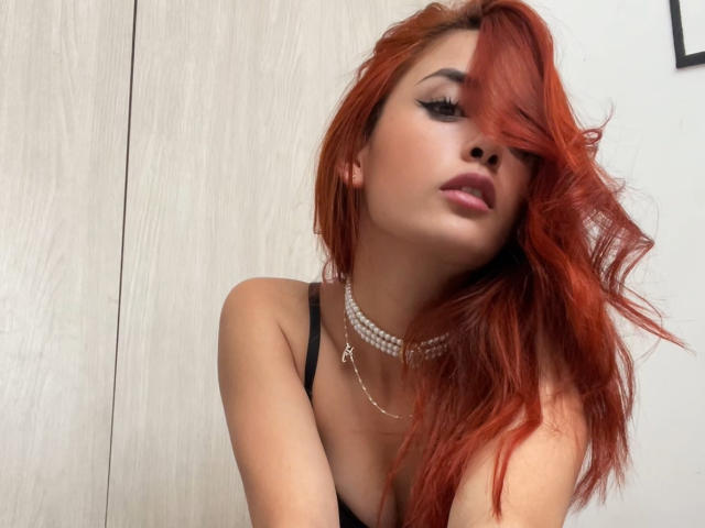KatherinGrey tem cabelo ruivo e ondulado que cobre um olho, delineador preto, lábios rosa e um colar de pérolas. Ela está vestindo uma blusa preta, que mostra seus seios..