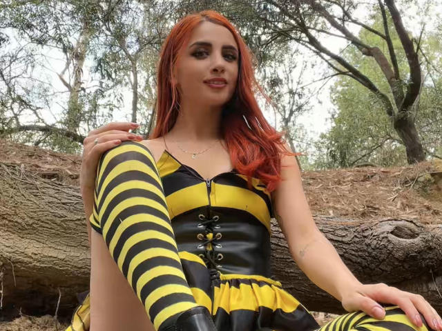 Katherin Grey, com cabelo ruivo, vestido listrado em amarelo e preto, e meias altas combinando, sentada em um tronco, sorrindo, mostrando sua perna e um corpete de couro com detalhes de cadarço..