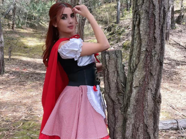 KatherinGrey with red hair, wearing a red cape, black corset, white puffy shirt, and red-checkered skirt, stands by a tree, touching her hair, with a playful smile.