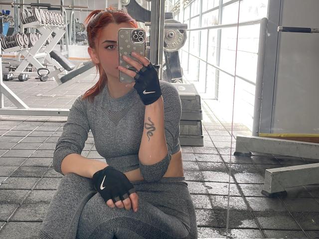 KatherinGrey with red hair, gray sports bra and pants, black Nike fingerless gloves, taking mirror selfie in gym, showing tattoo on right arm, holding iPhone.