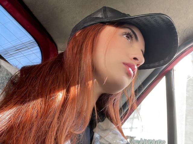 KatherinGrey with long red hair, wearing a black cap, looking up, sunlight on her face, glossy pink lips, soft brown eyes, and a plaid shirt, inside a car.
