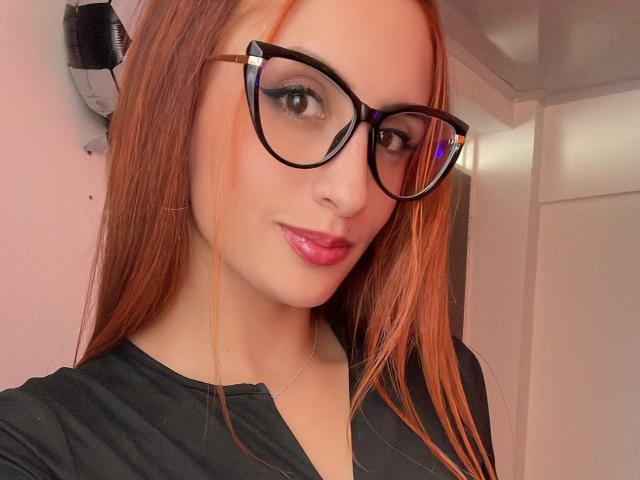 KatherinGrey with red hair, black glasses, pink lipstick, and black top, showing off her cute face and small nose, looking at the camera with a subtle smile.
