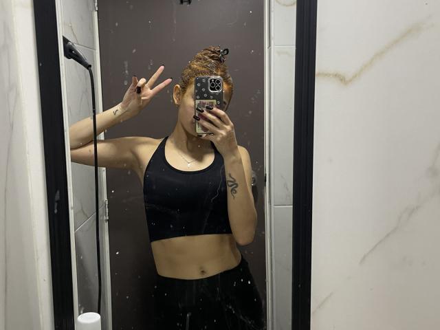 KatherinGrey with brown hair in a bun, wearing a black sports bra, showing a peace sign with her right hand, taking a mirror selfie in a bathroom.