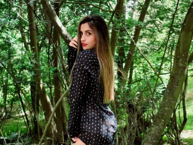 KatherinGrey with long, ombre hair, wearing a black polka dot shirt, looking over her shoulder with red lips, standing among green trees. Her ass is slightly visible.
