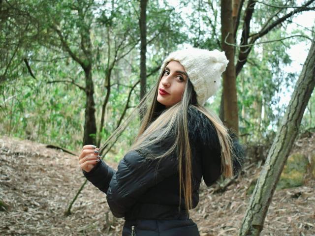 KatherinGrey with long brown hair, wearing a white knit beanie, black puffer jacket, and red lipstick, holding her hair in a forest. Boobs visible under jacket.