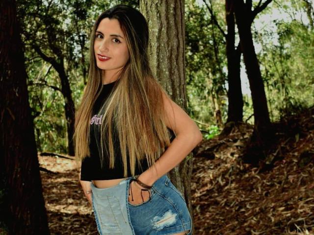 KatherinGrey with long blonde hair, black sleeveless top, and blue distressed jeans, standing in a forest, hands in pockets, smiling, showing her ass, brown tree trunk behind.