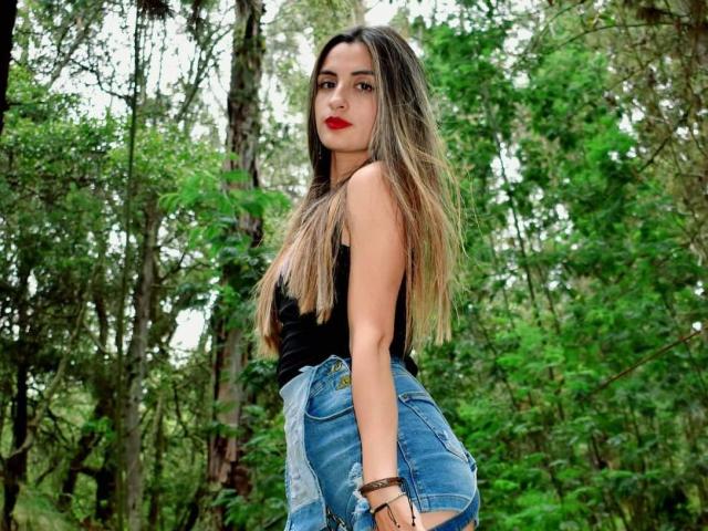 KatherinGrey with long blonde hair, red lipstick, black tank top, and high-waisted blue jeans, standing in a forest, looking over her shoulder, showing her ass.