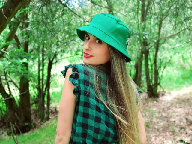 KatherinGrey, avec de longs cheveux blonds, un chapeau vert, du rouge à lèvres rouge et une chemise à carreaux verts, regardant par-dessus son épaule, debout dans une zone boisée. Les seins ne sont pas visibles..