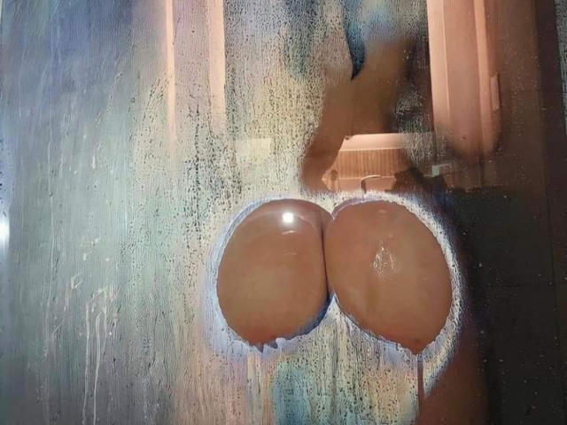 KatherinGrey's wet, round ass through a fogged-up shower door. Her brown butt cheeks are prominently displayed, with water droplets on the glass.
