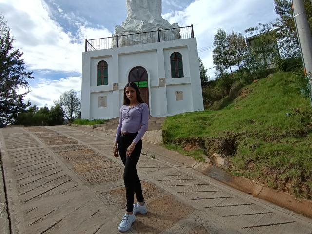 KatherinGrey with long brown hair, purple top, black pants, and white sneakers stands on a stone path, looking forward, in front of a white building with green windows.