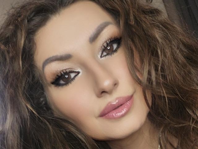 SweetAdrianax has wavy brown hair, dark eyebrows, glittery brown eyes with small white dots, and pink glossy lips. Her makeup is on point, looking sexy and hot.