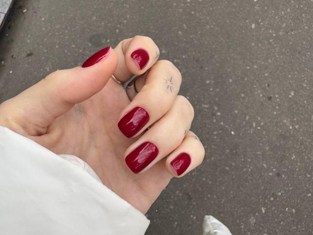 Tsukerberg's hand with shiny red nail polish, small star tattoo on ring finger, wearing a white sleeve, and a silver ring, standing on gray asphalt.