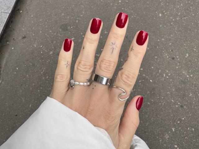 Tsukerberg's hand with shiny red nails, silver ring, and tiny cross tattoos on fingers, wearing a white sleeve, standing on a gray, textured surface.