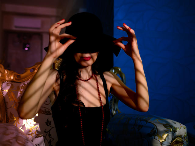 KamiLove with black hat, red beads, black top, slim arms, red nails, smiling, shadow stripes, holding hat, blue-patterned wall, ornate chair, soft lighting, red lipstick.