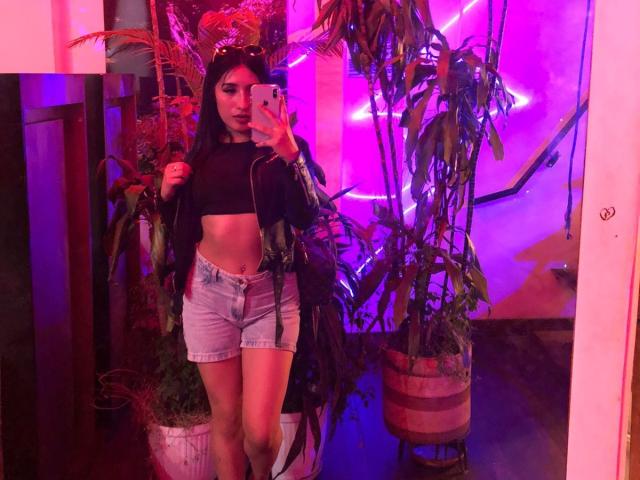 NickiClark with long dark hair, black crop top, showing abs, wearing high-waisted denim shorts, holding phone, standing in front of plants, purple neon lights, looking at mirror.