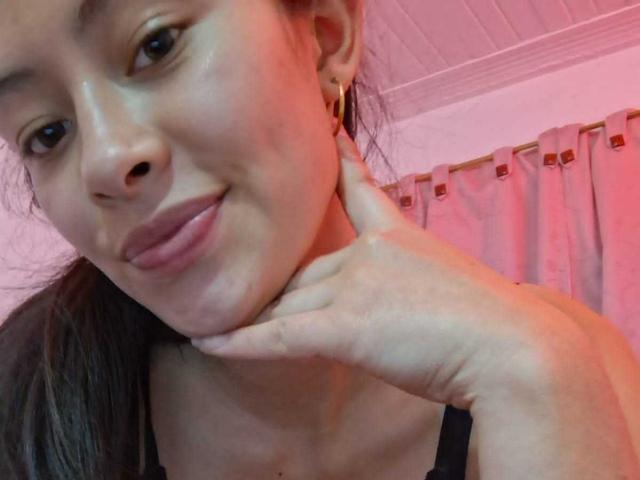 O rosto de TatianaRiosLove, com olhos castanhos, lábios rosados e pele bronzeada, segurando o queixo com a mão esquerda, usando brincos de ouro e uma blusa preta, em um quarto iluminado com luz rosa..