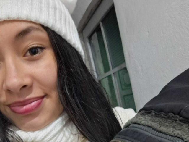 O rosto de TatianaRiosLove, com cabelos pretos longos, olhos castanhos, lábios rosados, um gorro de tricô branco e uma gola alta branca. Seios levemente visíveis sob uma jaqueta escura. Uma janela ao fundo..