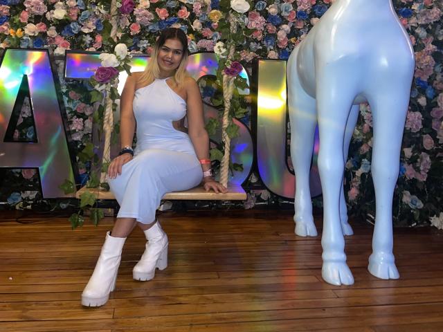 TarahDustin with big ass, white halter dress, white boots, sitting on wooden swing, floral wall, and blue horse statue, smiling. Blonde, pink wristbands, wooden floor.
