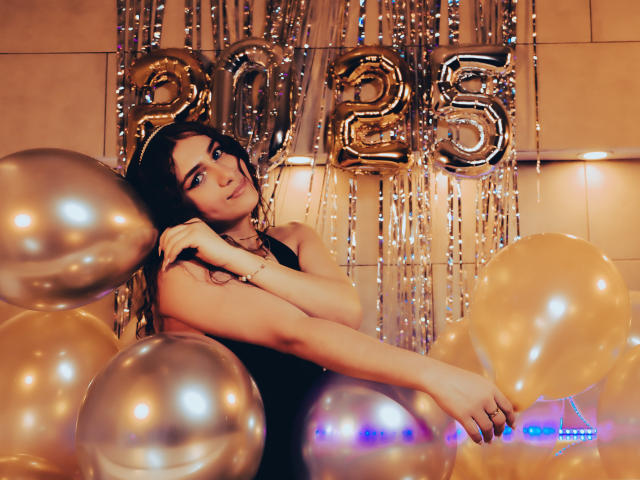 AndyDupont with long dark hair, wearing a black dress, holding gold balloons, arms crossed, in front of shiny silver numbers "25", looking slightly to the side with a serious expression.