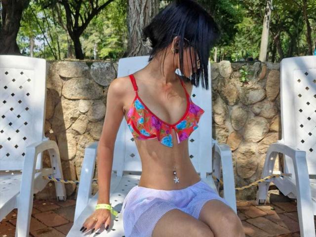 KylieGreenX with small tits, wearing a red, colorful floral bra, white mesh shorts, and a green wristband, sitting on a white chair, looking down, in a sunny outdoor setting.