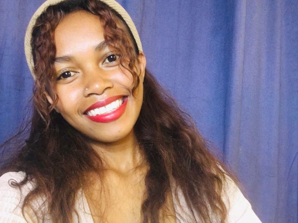BellaCeleste has dark brown curly hair, red lipstick, and is smiling with white teeth. She's wearing a beige hat and a white top against a blue backdrop.