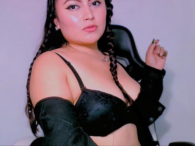 KristieJuteux with long braids, wearing a black bra, off-shoulder jacket, and pink cheek blush, showing her tits, in a chair, with a slight pout, and green nail.