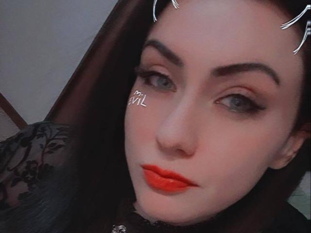 GoddessLiliMayhem has pale skin, dark eyebrows, blue eyes, red lipstick, and black floral lace top. She's wearing a nose ring and digital white heart sticker on her cheek.