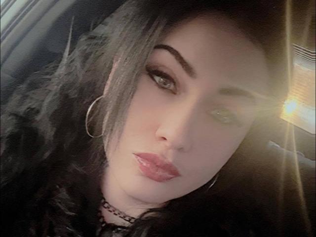 GoddessLiliMayhem has dark wavy hair, thick eyebrows, green eyes, glossy pink lips, a silver hoop earring, and a black choker. She's sitting in a car.