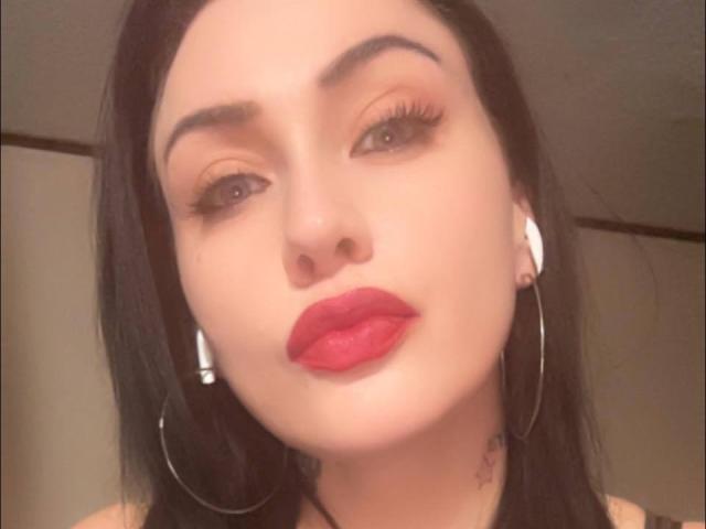 GoddessLiliMayhem has red lipstick, black hair, and is wearing white earbuds with large silver hoops, sporting thick eyebrows and long eyelashes, with a slightly pouty expression.